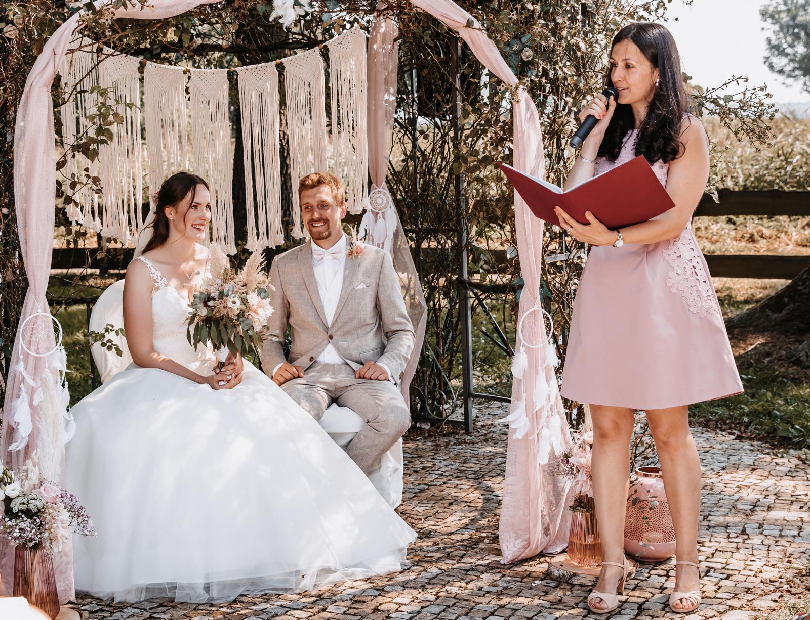 Magische freie Trauung in Brandenburg. Hochzeitspaar sitzt unter dem Bogen und lächelt, Traurednerin hält Mikro in der Hand